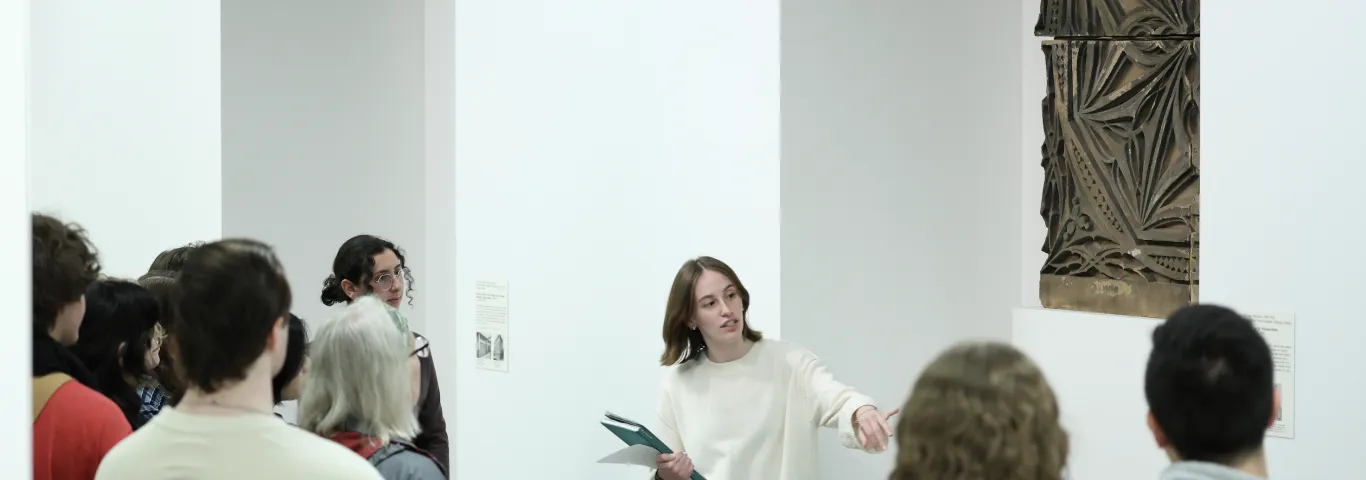 "Student leading tour stop in a gallery with a sculptural brown work hung on the wall"